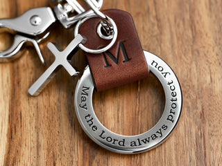 A custom brown leather keyring with a stainless steel ring and a cross charm, featuring engraved text on the ring.